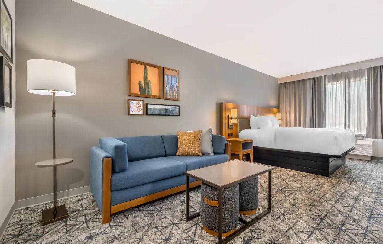 A hotel room with a blue sofa, a small table, a bed, a floor lamp, and artwork on the wall.