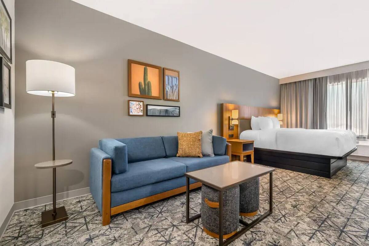 A hotel room with a blue sofa, a small table, a bed, a floor lamp, and artwork on the wall.
