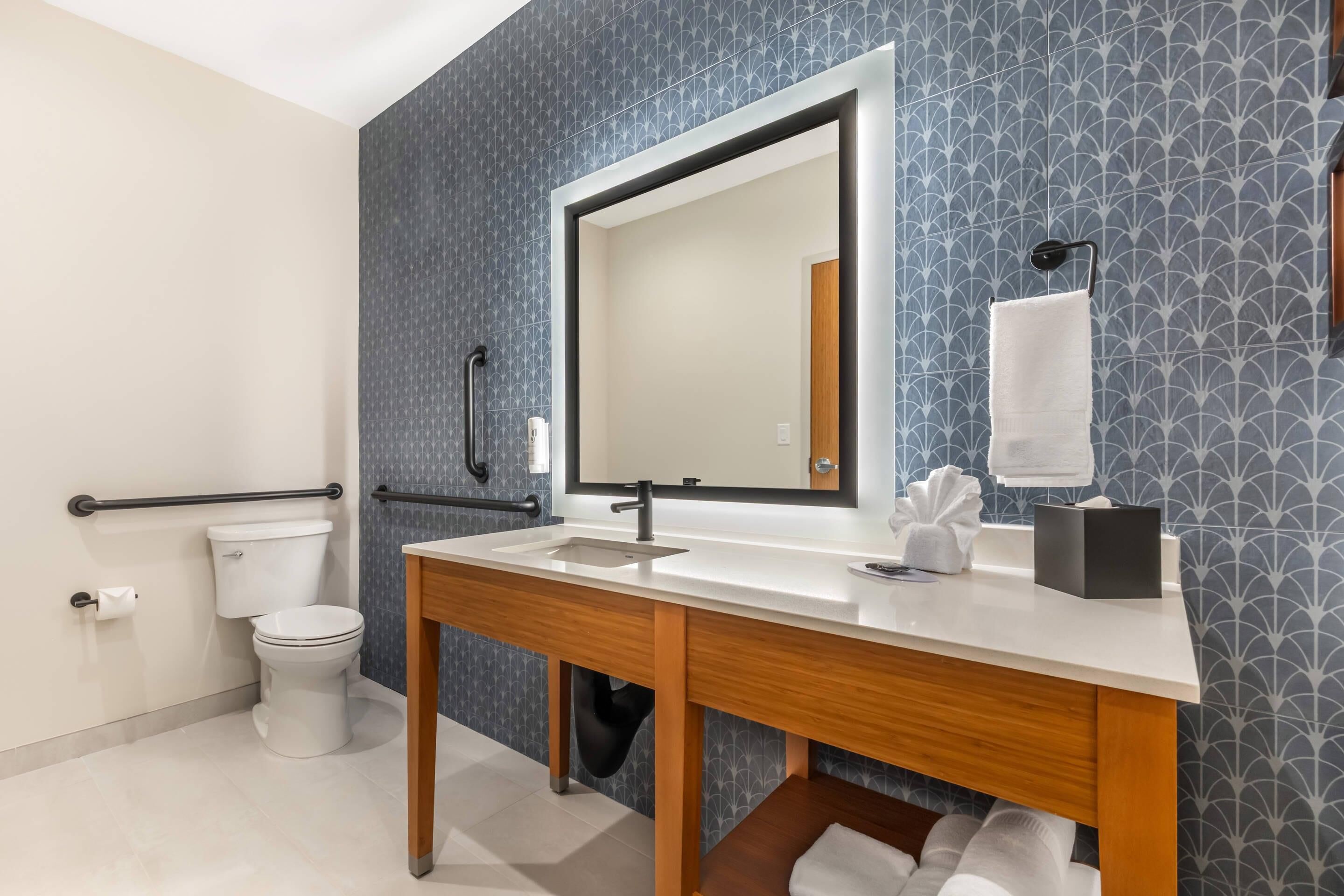 Un baño moderno con lavamanos, espejo y retrete blancos, tocador de madera y azulejos con diseños.