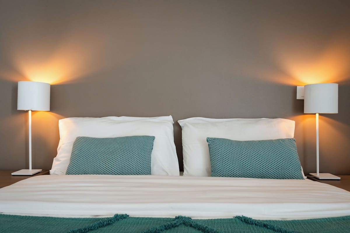A double bed with white linens and teal pillows, flanked by two wall-mounted lamps.