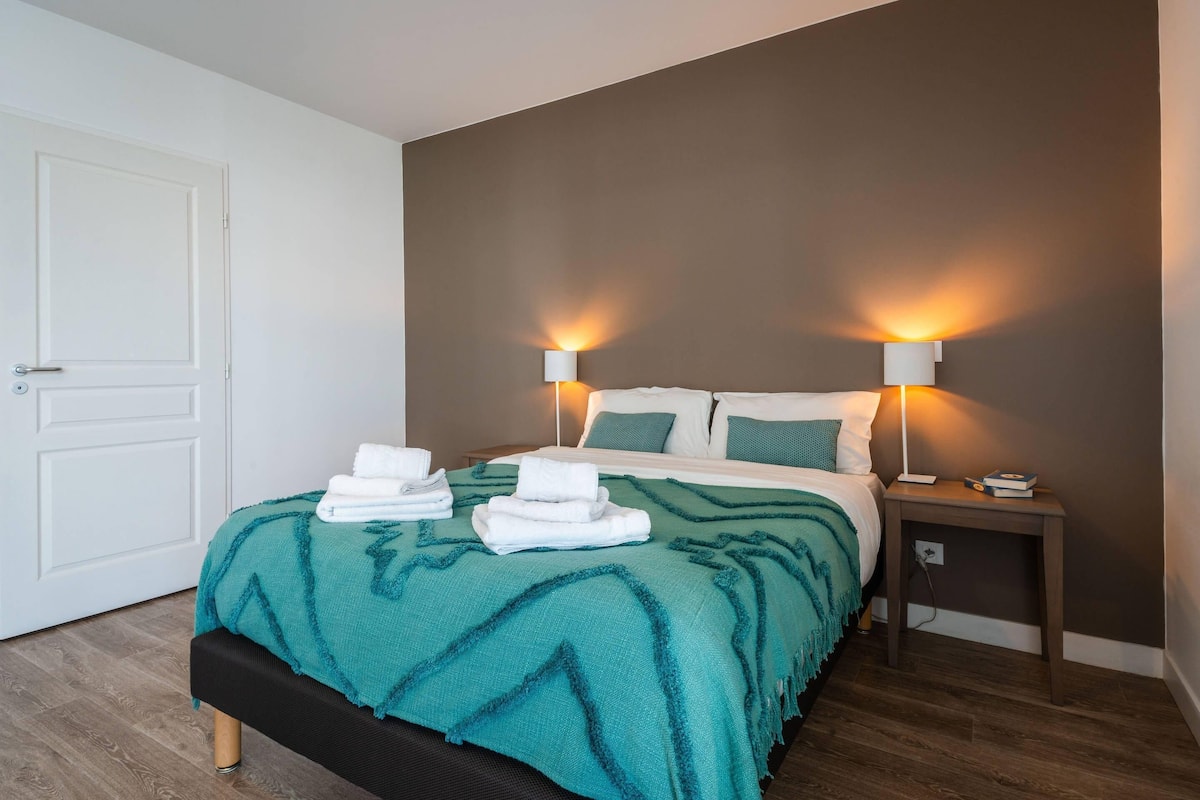 A neatly made bed with a teal blanket, two bedside lamps, and a small bedside table with a book and a phone.