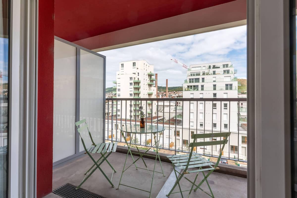 A balcony with two green chairs and a small table, overlooking a residential area with buildings and a construction crane.