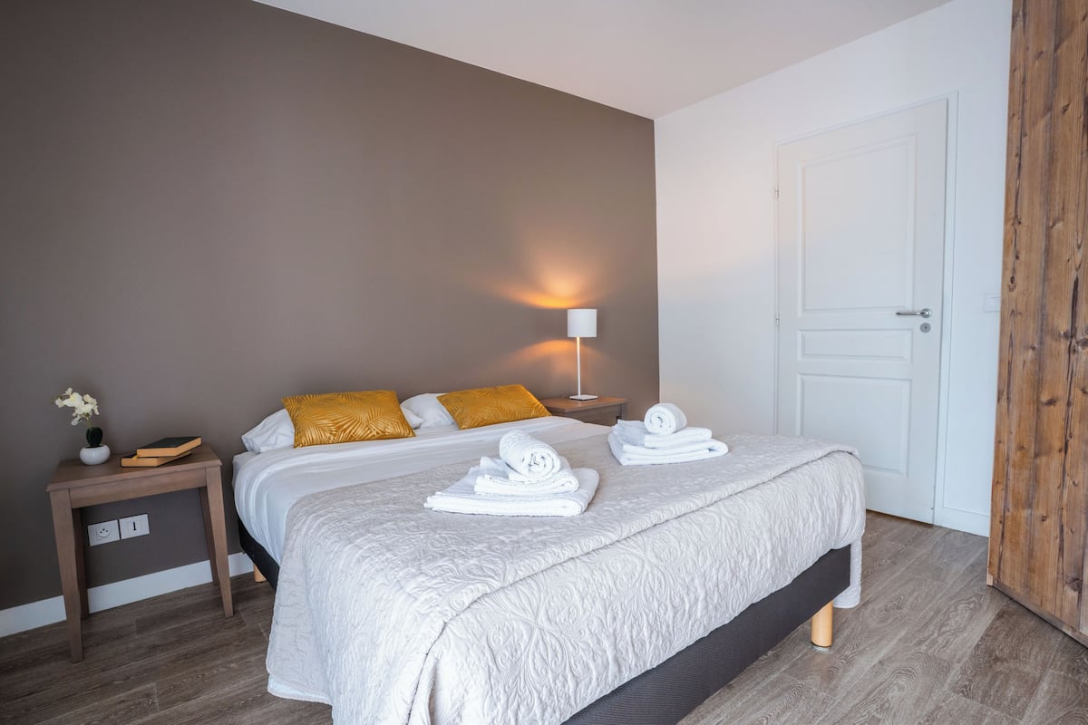 A neatly made bed with white linens, a bedside lamp, and a small table with a plant and books.