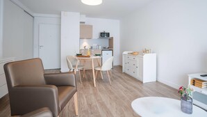 Una sala moderna con un sofá de cuero marrón, una mesa de centro redonda y un comedor con mesa y sillas blancas. Se ve una cocina con armarios blancos y un microondas al fondo.