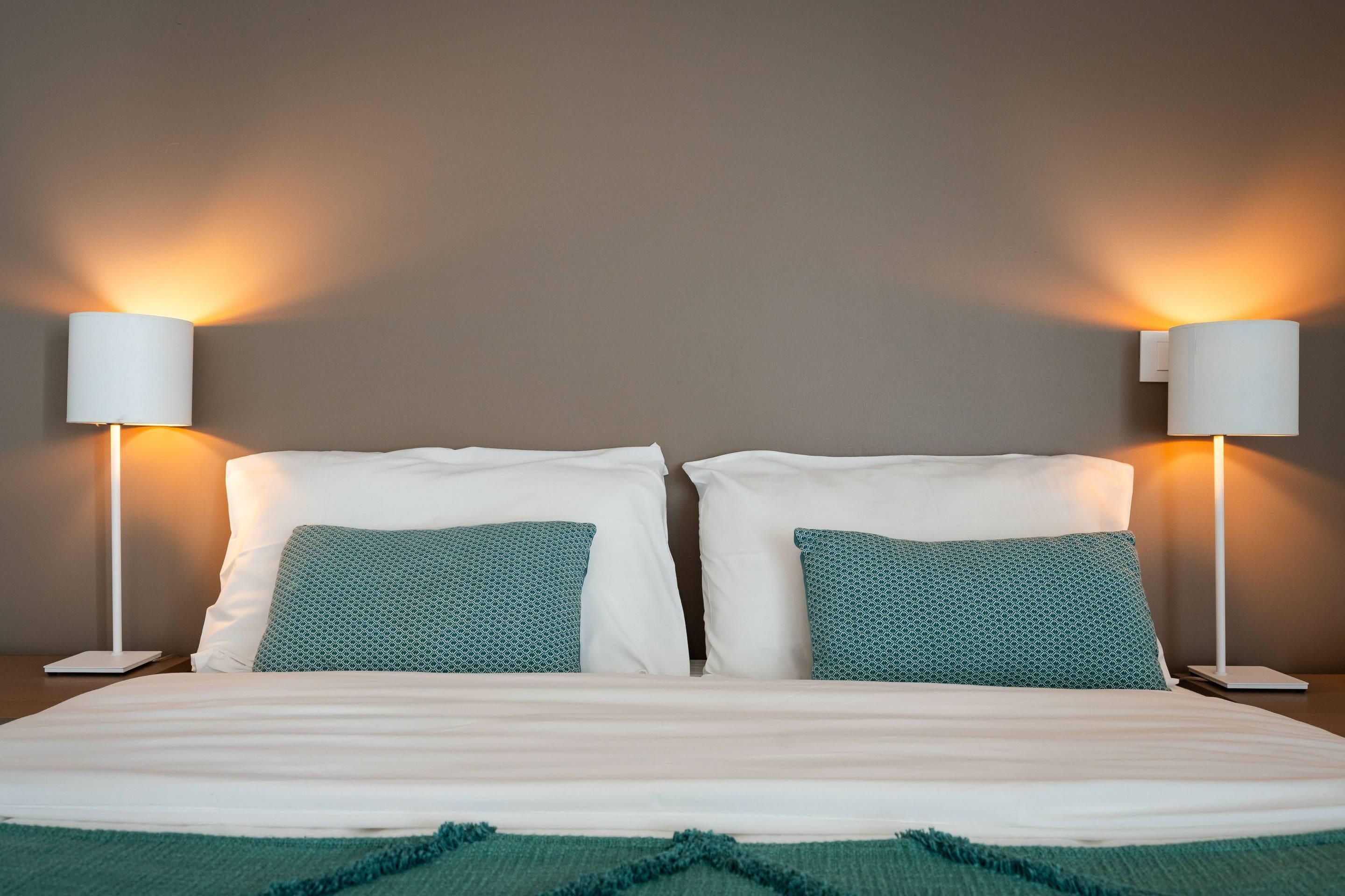 A neatly made bed with white linens and two green patterned pillows, flanked by two bedside tables with white lamps.