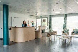 Uma área de recepção com uma mesa de madeira curva, duas poltronas, uma mesa de centro e cortinas estampadas.