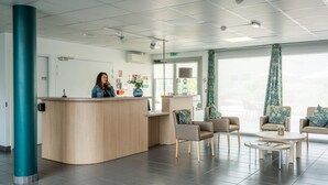 Una zona de recepción con un escritorio de madera curvo, dos sillones, una mesa de centro y cortinas con estampado.