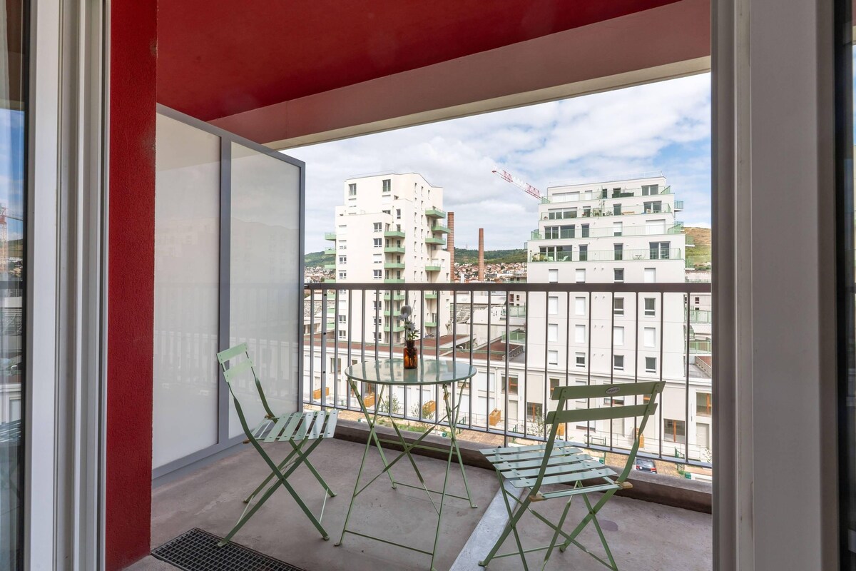 Uma varanda com duas cadeiras verdes e uma mesa pequena, com vista para uma área residencial com edifícios e uma grua de construção.