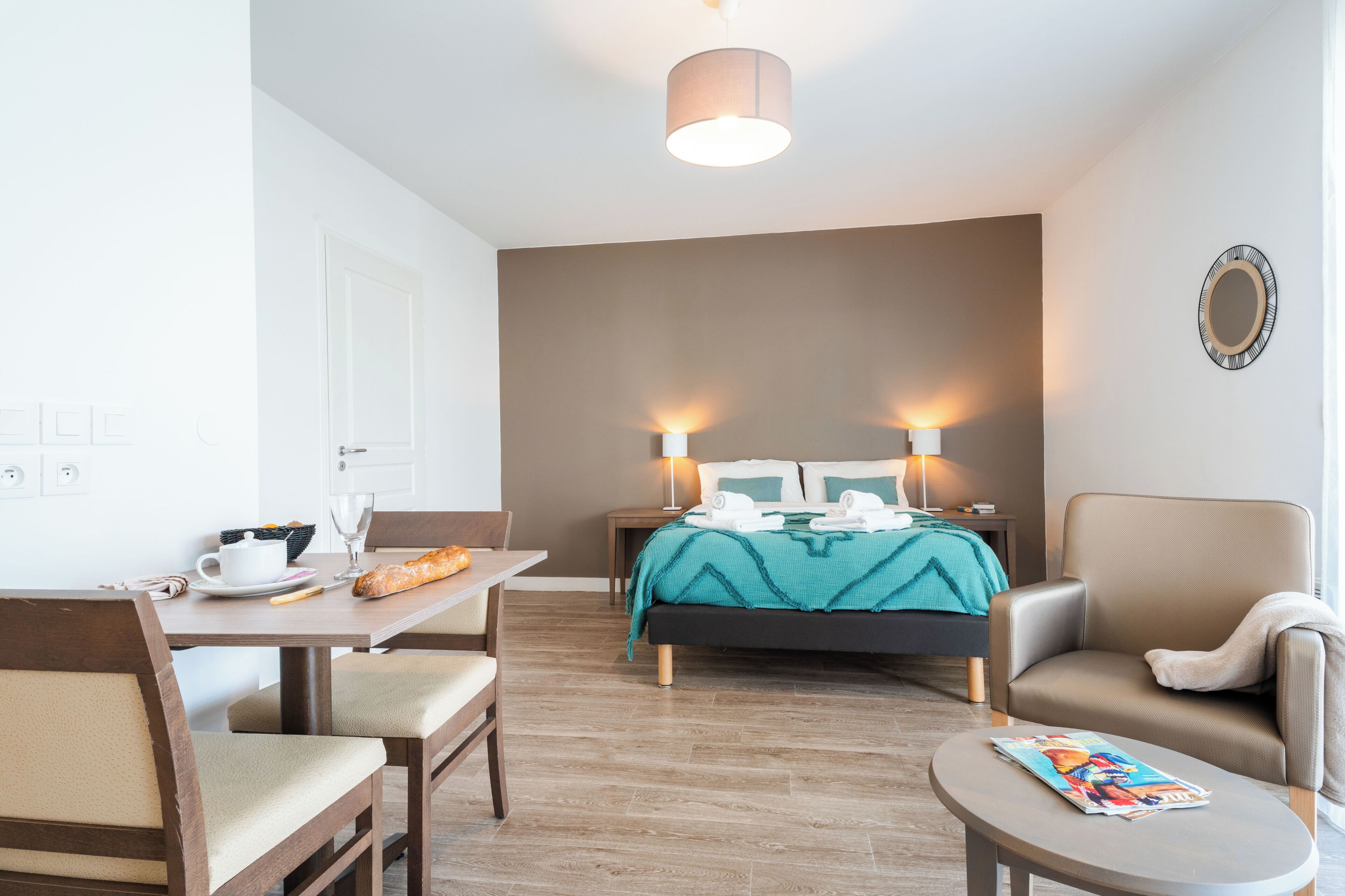 A hotel room with a bed, two bedside lamps, a table with a chair, a small round table, and a magazine on the small table.
