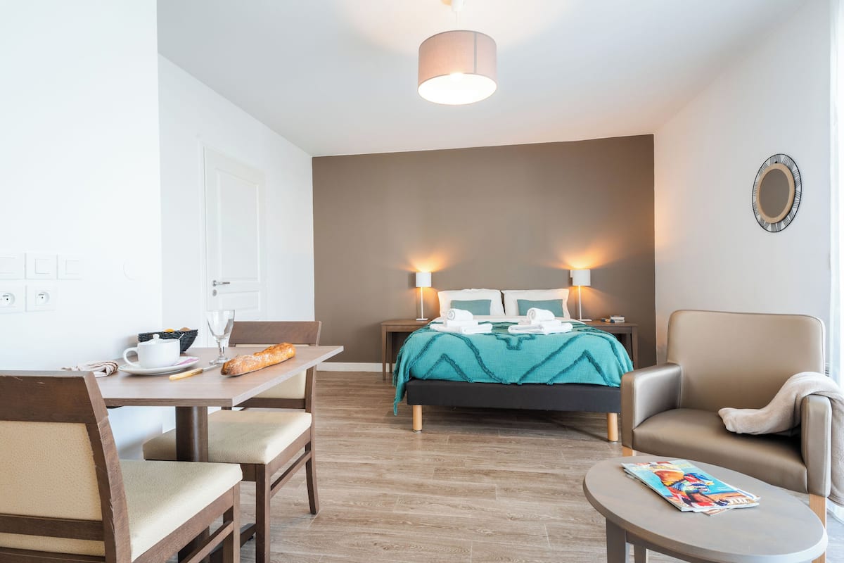 A hotel room with a bed, two bedside lamps, a table with a chair, a small round table, and a magazine on the small table.