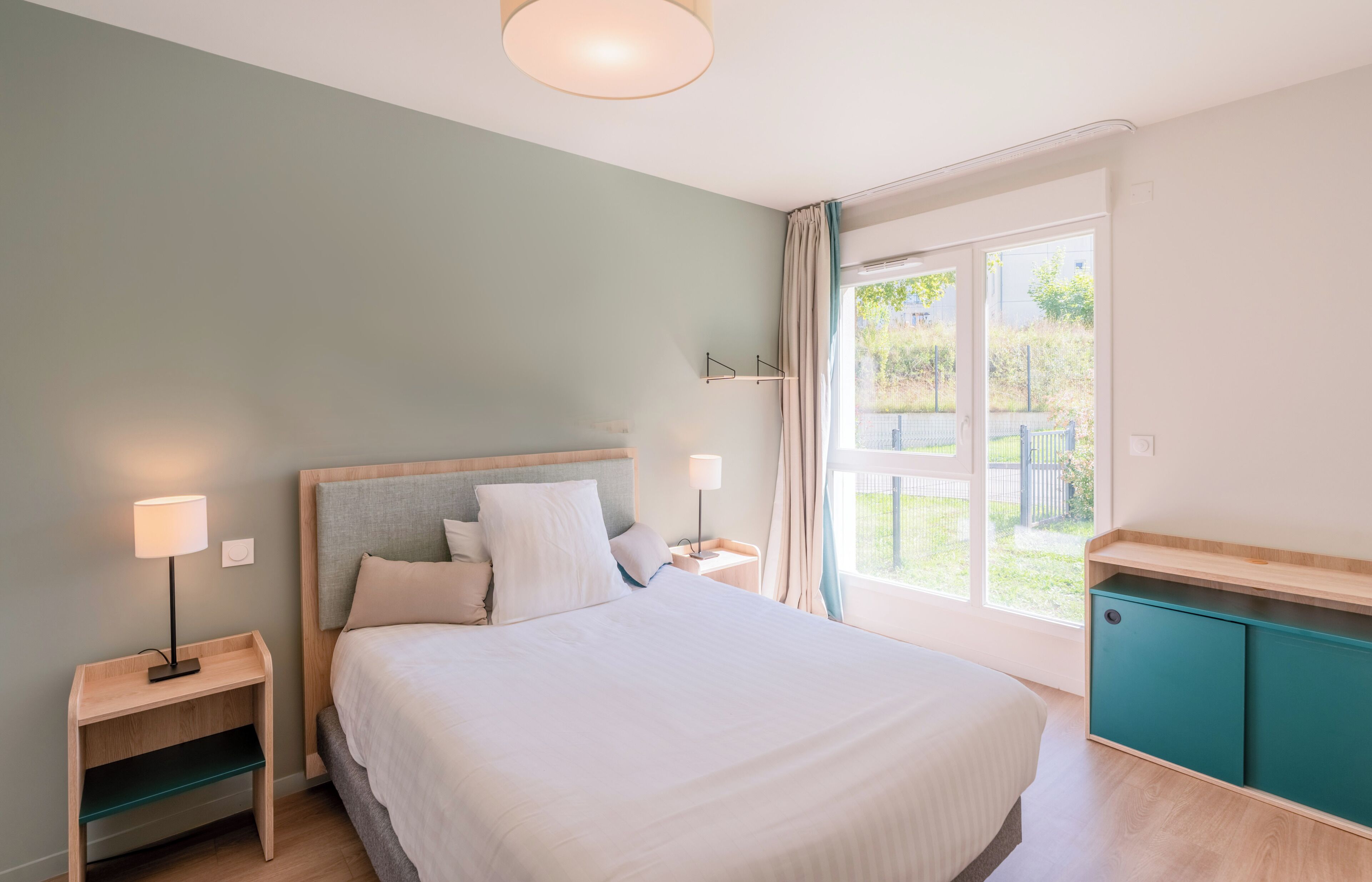 A bedroom with a bed, bedside tables, a nightstand, a window with curtains, and a view of the outdoors.