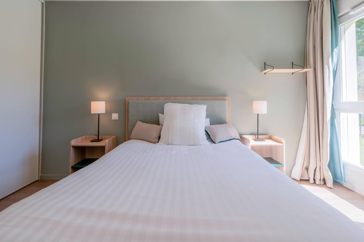 A neatly made bed with a white comforter and pillows, two bedside tables with lamps, a headboard, and a window with curtains.