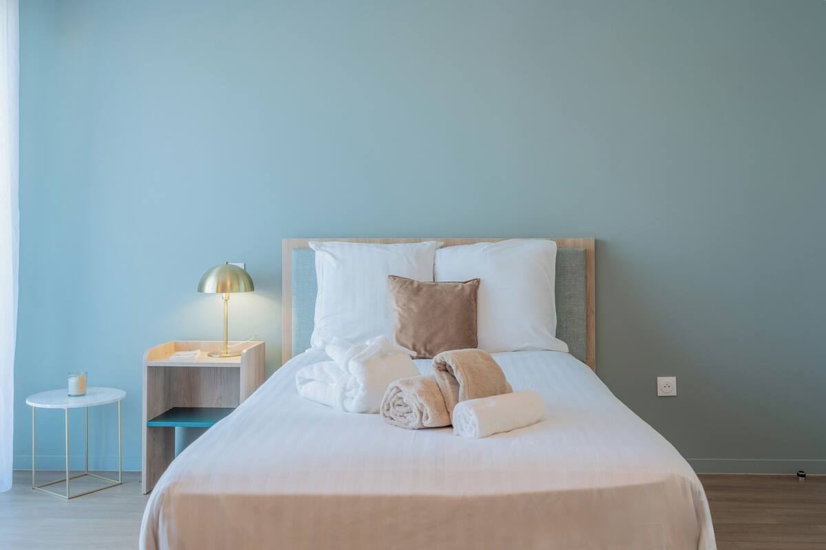 A neatly made bed with white linens and rolled towels, a bedside table with a lamp, and a small round table with a candle.