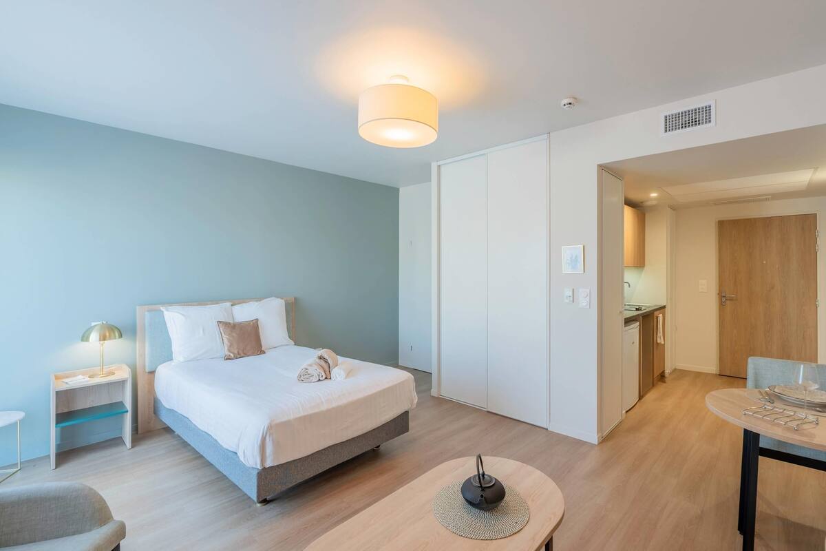 A modern hotel room with a bed, bedside table, lamp, and a dining area with a table and chairs.