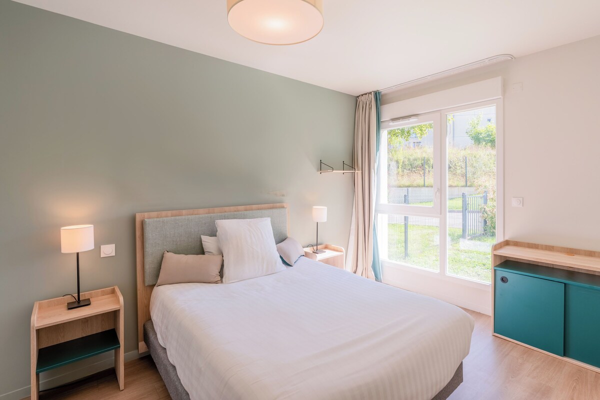 A bedroom with a bed, bedside tables, a nightstand, a window with curtains, and a view of the outdoors.