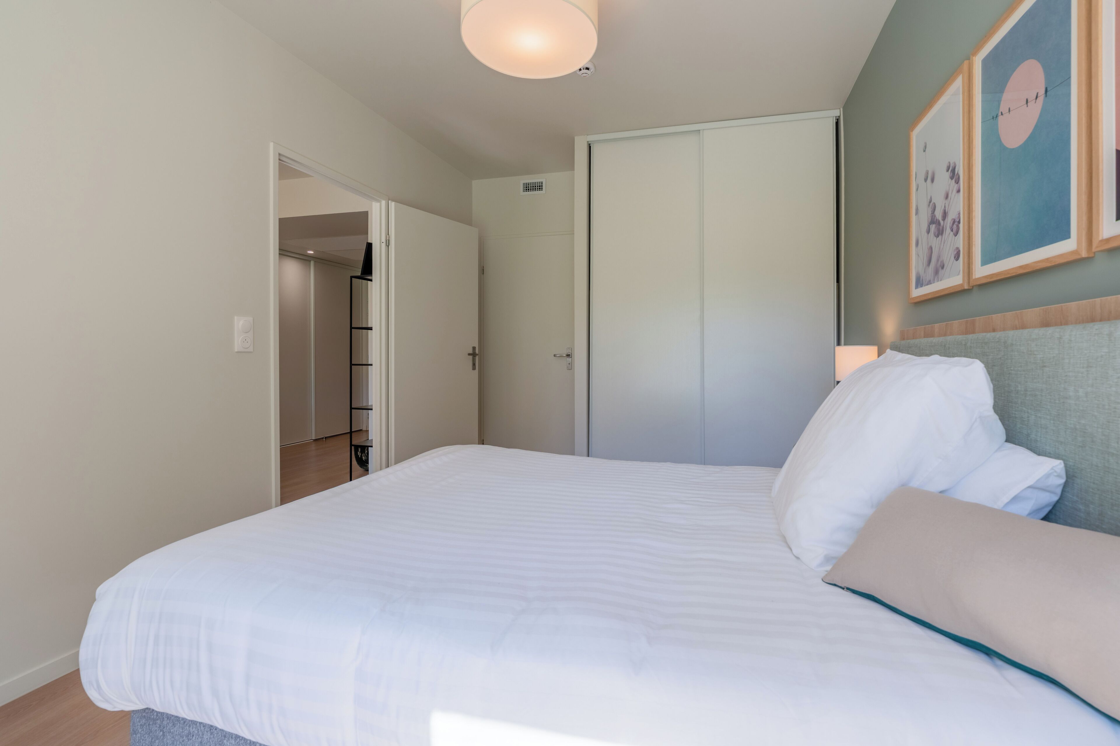 A neatly made bed with white bedding, a grey headboard, and a beige pillow. There is a door leading to a hallway with a white door and a light switch on the wall.