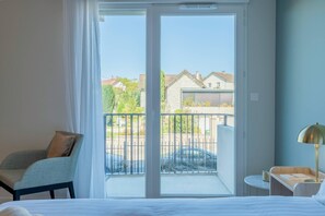 Ein Hotelzimmer mit einem Bett, einem Sessel, einem kleinen Tisch mit Lampe und einem Balkon mit Blick auf Häuser und einen Zaun.
