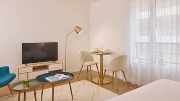 Une chambre d’hôtel comprenant un lit, une télévision, un bureau avec une lampe, deux chaises et une fenêtre avec des rideaux légers.