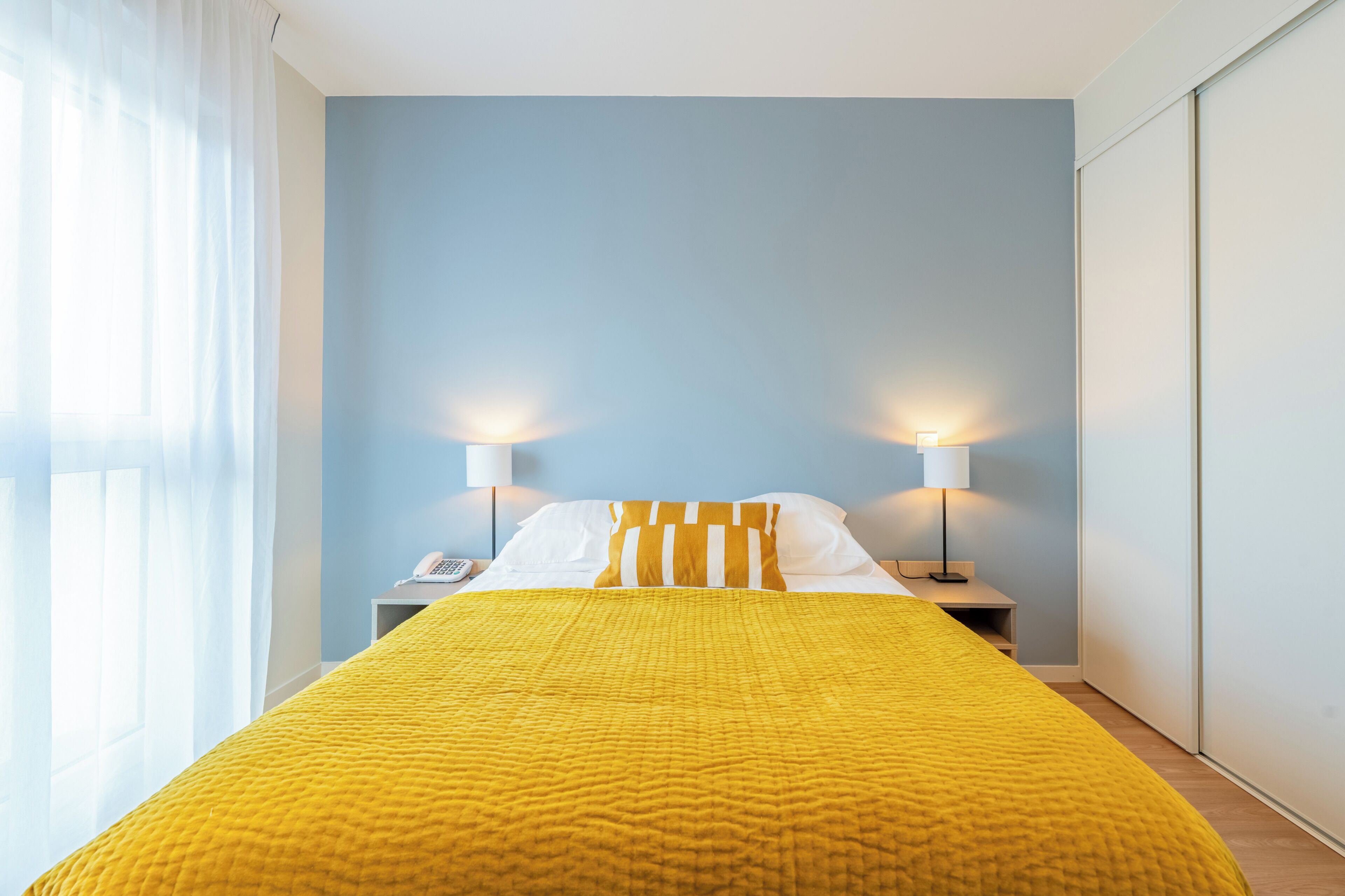 A hotel room with a bed, two bedside lamps, a telephone, and a window with sheer curtains.
