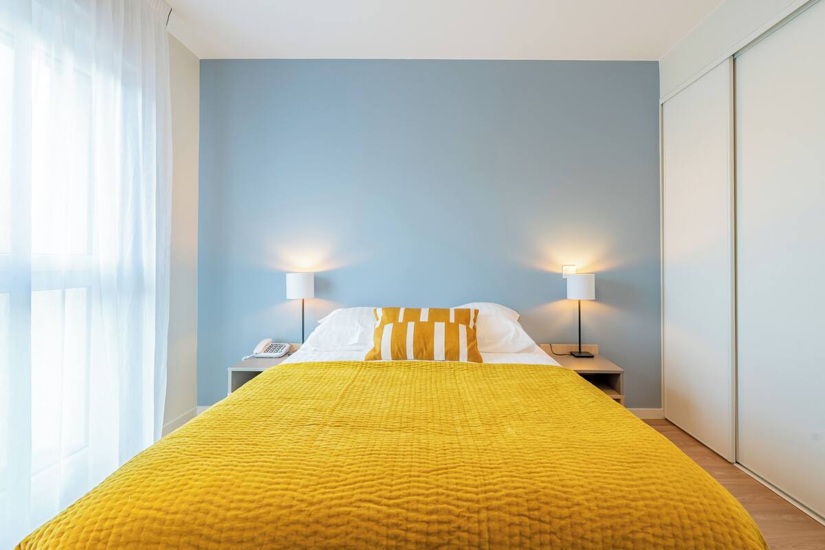 Una camera d'albergo con un letto, due lampade da comodino, un telefono e una finestra con tende leggere.