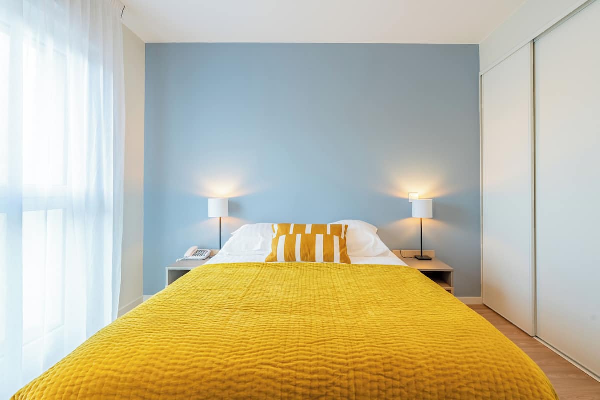 Una habitación de hotel con una cama, dos lámparas de noche, un teléfono y una ventana con cortinas traslúcidas.