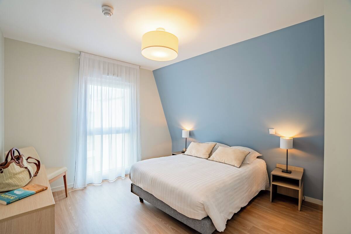 Una camera da letto con un letto, lampade da comodino, una sedia, un tavolo e una finestra con tende.