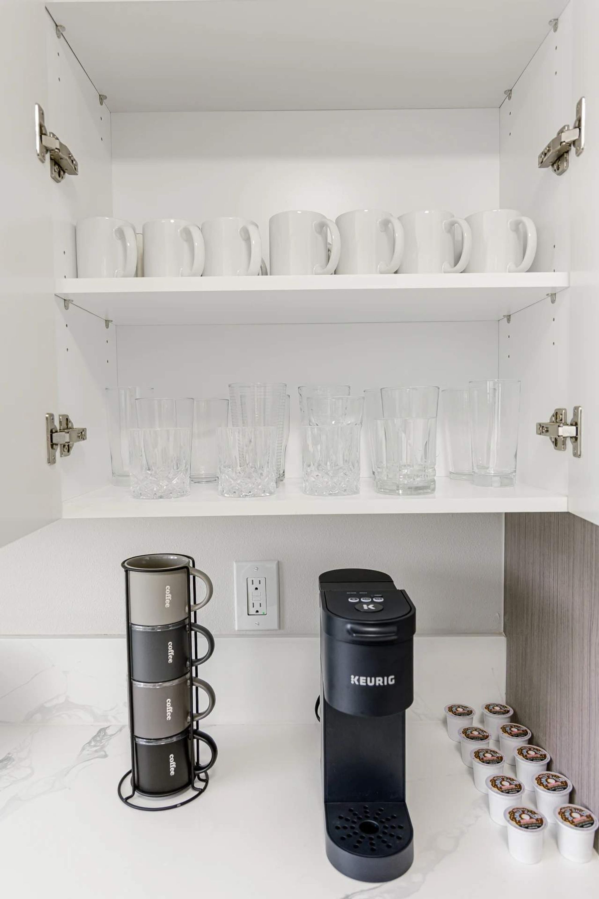 Une cuisine avec des étagères blanches où sont posées des tasses et des verres transparents. En dessous, on peut voir une machine à café Keurig noire et un présentoir à tasses contenant plusieurs tasses.