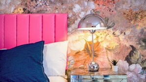 A bedroom with a pink headboard, a white bed, a blue pillow, a glass nightstand with a lamp, and a floral wall mural. - Comfortable Studio with Great City Access (Sheffield)