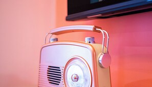 Una radio antigua con asa, apoyada contra una pared rosa con un televisor encima.