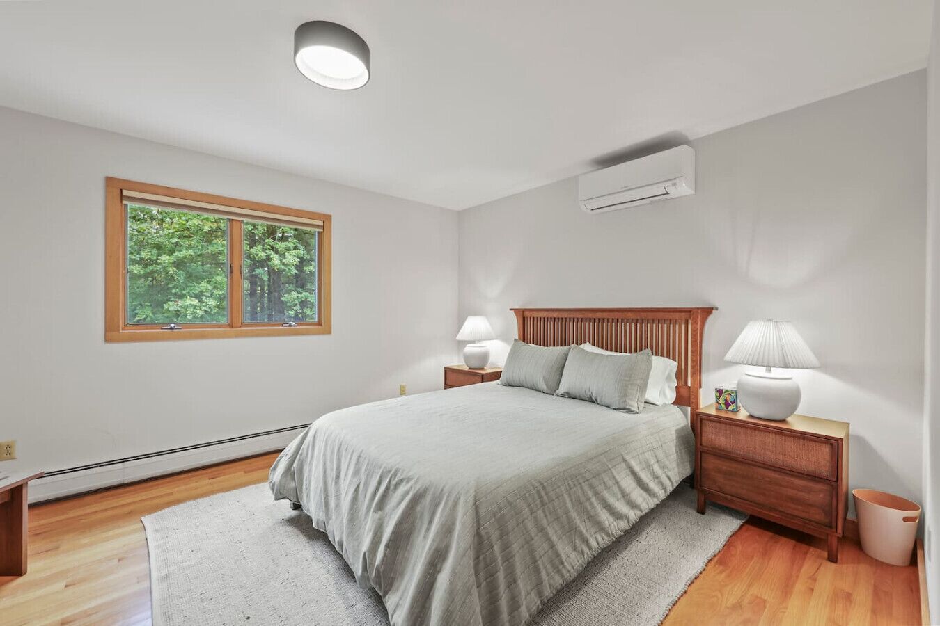 Een slaapkamer met een groot bed, een houten hoofdbord, twee nachtkastjes met lampen, een raam met uitzicht op bomen en een airconditioner aan de muur.