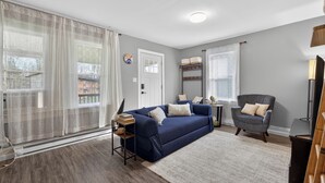 Een woonkamer met een blauwe bank, een grijze fauteuil, een klein tafeltje en een groot raam met gordijnen.