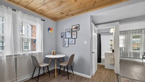 Een kamer met een houten plafond, een eettafel met stoelen, een kleine witte tafel en een deur die naar een andere kamer leidt.
