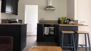 Cocina moderna con armarios oscuros, campana extractora de acero inoxidable y horno empotrado. Hay dos taburetes y una mesa de madera.