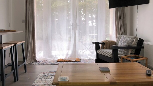 Una sala de estar con un sofá, una mesa de centro, un televisor montado en la pared y un ventanal corredizo con cortinas.