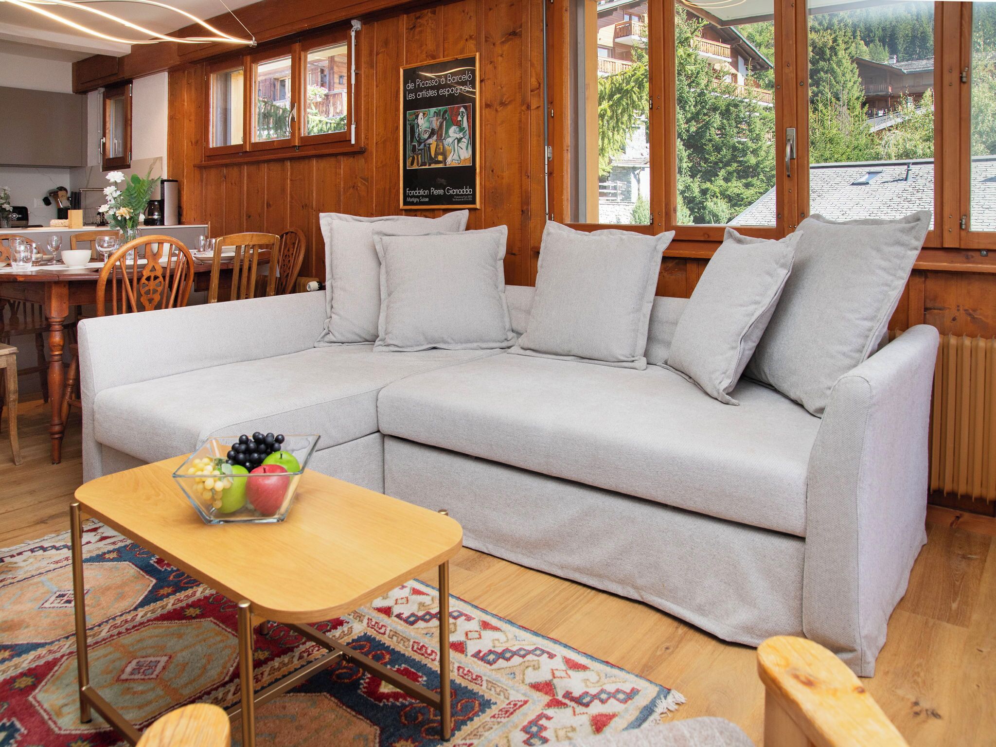 Un salon chaleureux avec un canapé gris, une petite table en bois sur laquelle sont disposés des fruits, et une vue sur un paysage montagneux grâce à de grandes fenêtres.