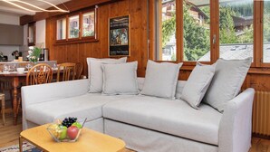 A cozy living room with a grey sofa, a small wooden table with fruit, and a view of a mountainous landscape through large windows.