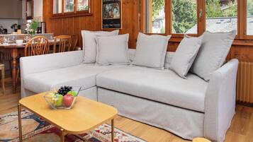 Un salon chaleureux avec un canapé gris, une petite table en bois sur laquelle sont disposés des fruits, et une vue sur un paysage montagneux grâce à de grandes fenêtres.
