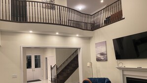 Ein großzügiges Wohnzimmer mit hoher Decke, einer Treppe zu einer oberen Ebene, einem flachbildschirmfernseher über einem Kamin und einem blauen Sessel.