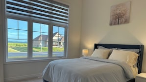 Ein Schlafzimmer mit großem Fenster, einem Bett mit weißer Bettwäsche, einem Nachttisch mit Lampe und einem Wandbild.