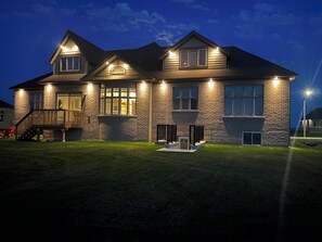 Une maison à deux étages avec une façade en pierre, une terrasse à l’avant et un éclairage extérieur soigné.