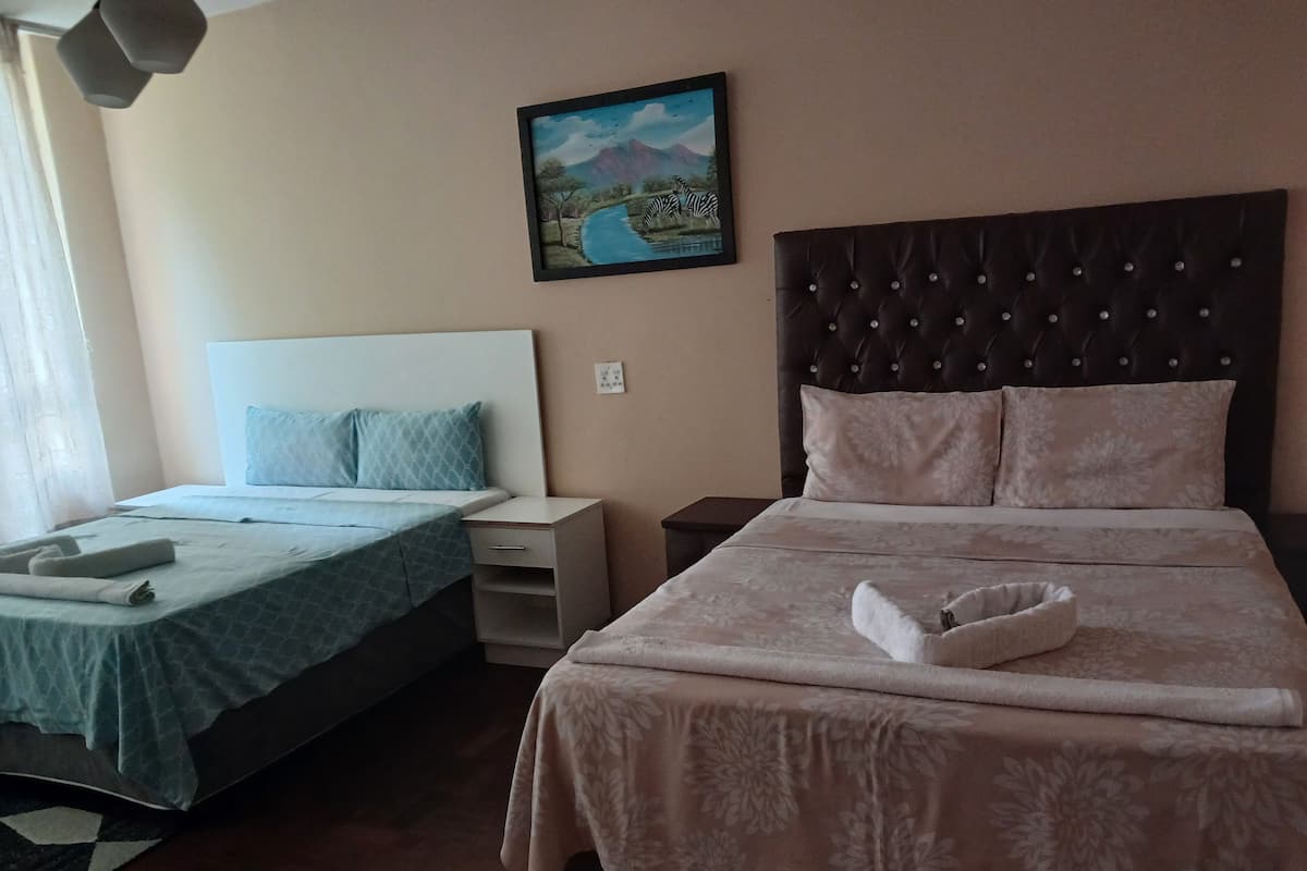 A hotel room with two beds, a nightstand, a bedside table, a painting on the wall, and a window with curtains.
