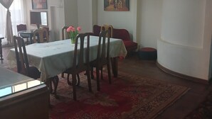 Ένας χώρος τραπεζαρίας με ένα τραπέζι στρωμένο για τέσσερα άτομα, το οποίο περιβάλλεται από καρέκλες. Στο πάτωμα υπάρχει ένα κόκκινο χαλί και στον τοίχο διακρίνεται ένας πίνακας.