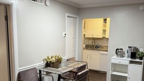 A compact kitchen with a dining area, featuring a glass-top table, metal chairs, and a white cabinet with a built-in oven. - Quiet Corner ATL (Atlanta)