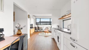 A modern kitchen with a washing machine, a dining area with a round table, and a view of the outdoors through a large window. - The Jitterbug - Sleeping 4 Guests (The Hill)