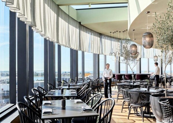 Ein Restaurant mit hohen Decken, großen Fenstern und Blick auf die Skyline. Tische sind für das Essen gedeckt, und das Personal ist anwesend.