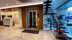 A modern hotel lobby with a reception desk, a glass elevator, a staircase with a view of a pool, and a sign indicating a rooftop terrace. - THE PARADISE PLATINUM (Kanchipuram)