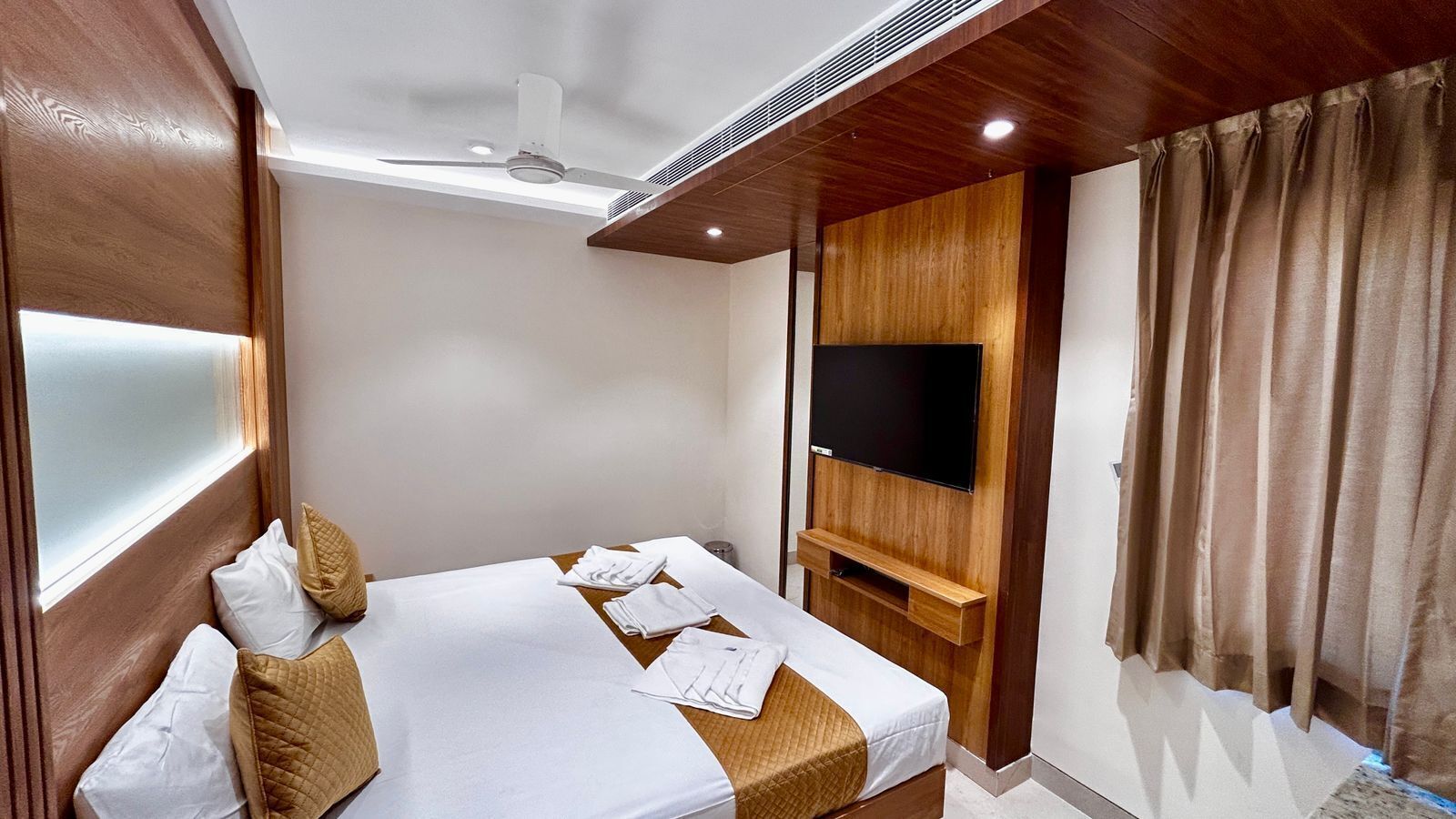 Habitación de hotel con cama, televisor de pantalla plana montado en la pared y ventana con cortinas.