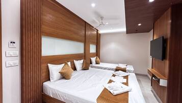 A hotel room with a large bed, two smaller beds, wooden paneling, a ceiling fan, a flat-screen TV, and a wall-mounted shelf.