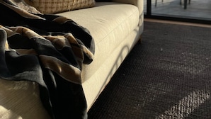 Ein Sofa mit einem gewebten Kissen und einer dunklen Decke, vor einem großen Fenster mit Blick auf Bäume und eine Brücke.