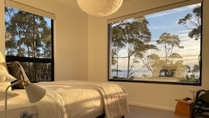 Ein Schlafzimmer mit einem großen Bett, einer Nachttischlampe, einem kleinen Tisch mit einer Pflanze und Blick auf Bäume durch ein Fenster.